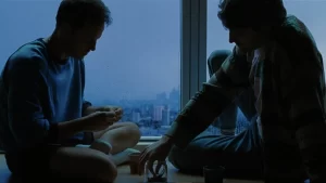 Adam and Harry sit on a floor in front of a large window with an urban view. Its dimly lit, suggesting dawn or dusk. One person wears a sweater and shorts, while the other wears a jacket and jeans. They appear to be having a casual conversation or drink.