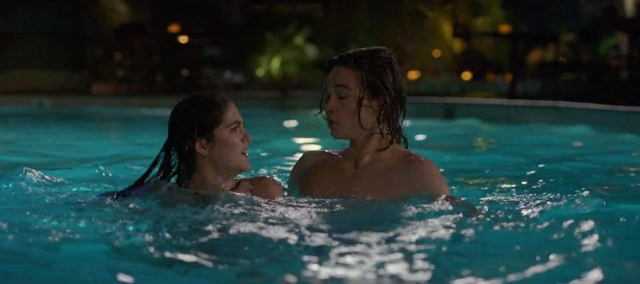 Two people are standing in a swimming pool at night. The woman on the left is smiling, while the man on the right has a neutral expression. The pool is surrounded by dimly lit trees and plants in the background.