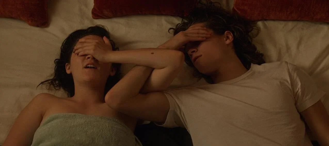 Two people lying on bed, covering their faces.