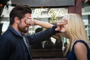 A man and a woman stand facing each other outside a cafe. Each is playfully covering the others eyes with their hand. The cafes sign is partially visible in the background.