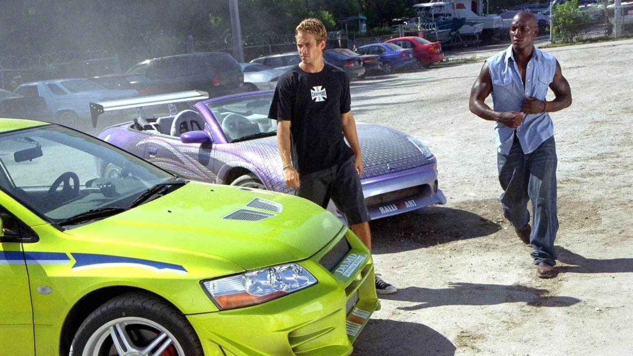 Two men stand next to a bright green car. Another car is parked behind them in a sunny, open area with other vehicles in the background.