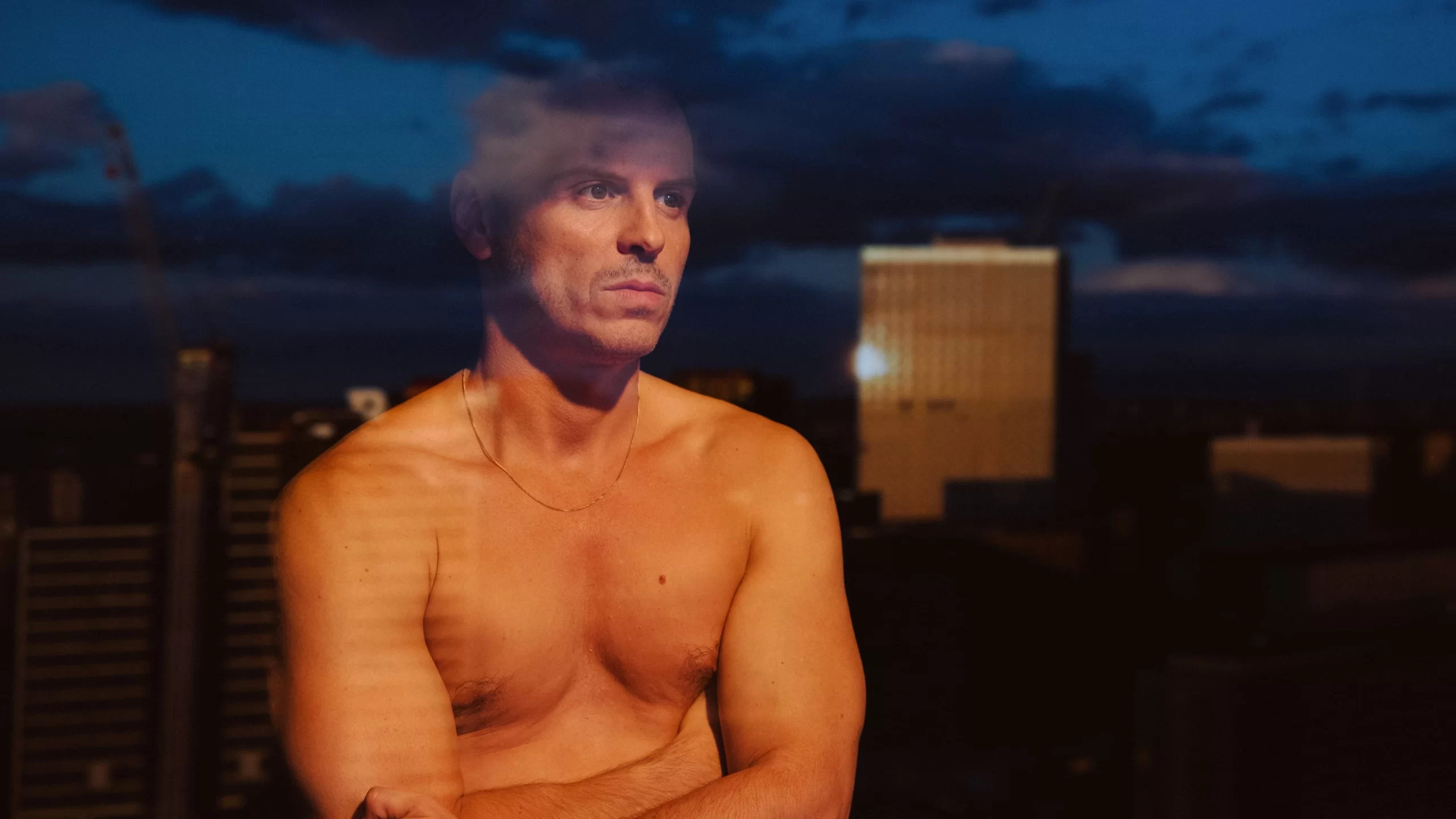 A shirtless Adam, embodying the solitude of All of Us Strangers, sits with arms crossed, gazing out a window in the evening.