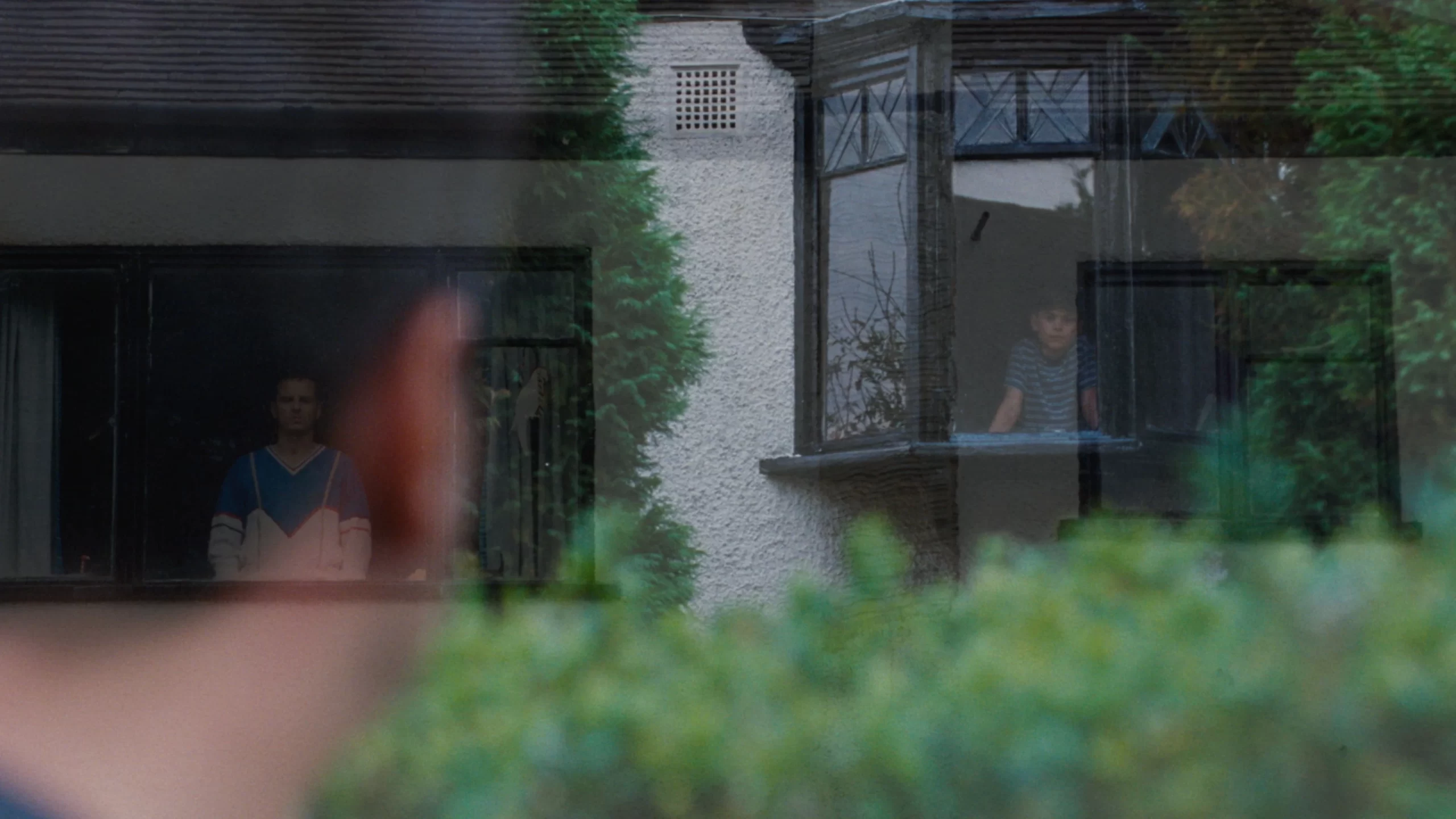 Adam is observing a house through a window, with two people visible inside. One is sitting by a bay window, while the other stands by a different window.
