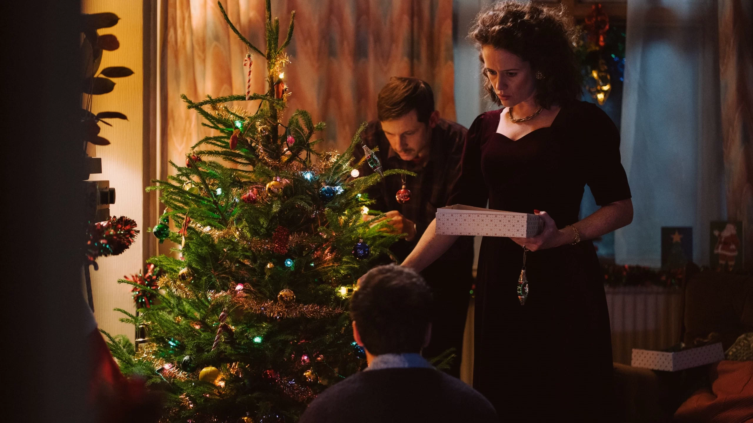 Adam's parents decorate a Christmas tree adorned with colourful lights and ornaments. The woman holds a white box with red polka dots. The room is dimly lit, creating a warm, festive atmosphere.