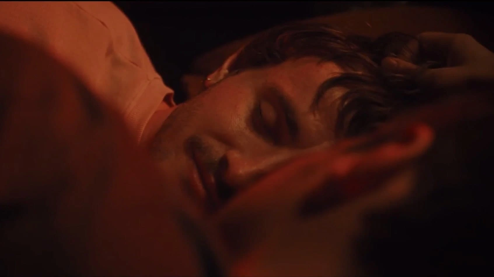Two people lie close together in a dimly lit setting, reminiscent of a scene from All Of Us Strangers. One, with short hair and facial hair, rests gently against the partially visible other. The warm lighting creates a cozy atmosphere.