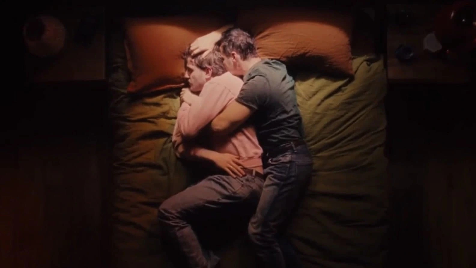 Two people lie on a bed under a green blanket and brown pillows. One, in a pink shirt and gray pants, rests on their side while the other, wearing a dark t-shirt and jeans, lovingly embraces them from behind—as if they were characters straight out of All Of Us Strangers.