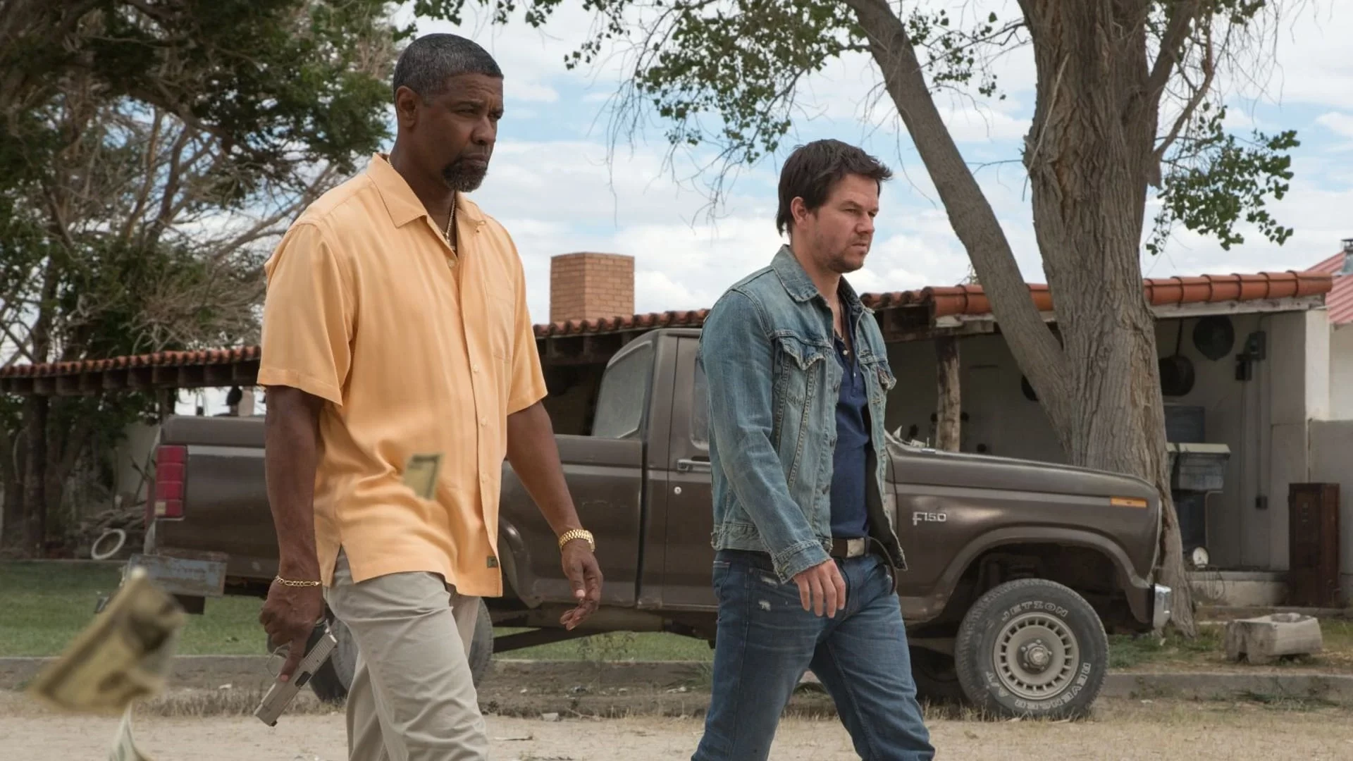 Two men walk outdoors beside a pickup truck. One wears a light orange shirt and holds a gun, the other wears a denim jacket. Trees and a building are in the background.