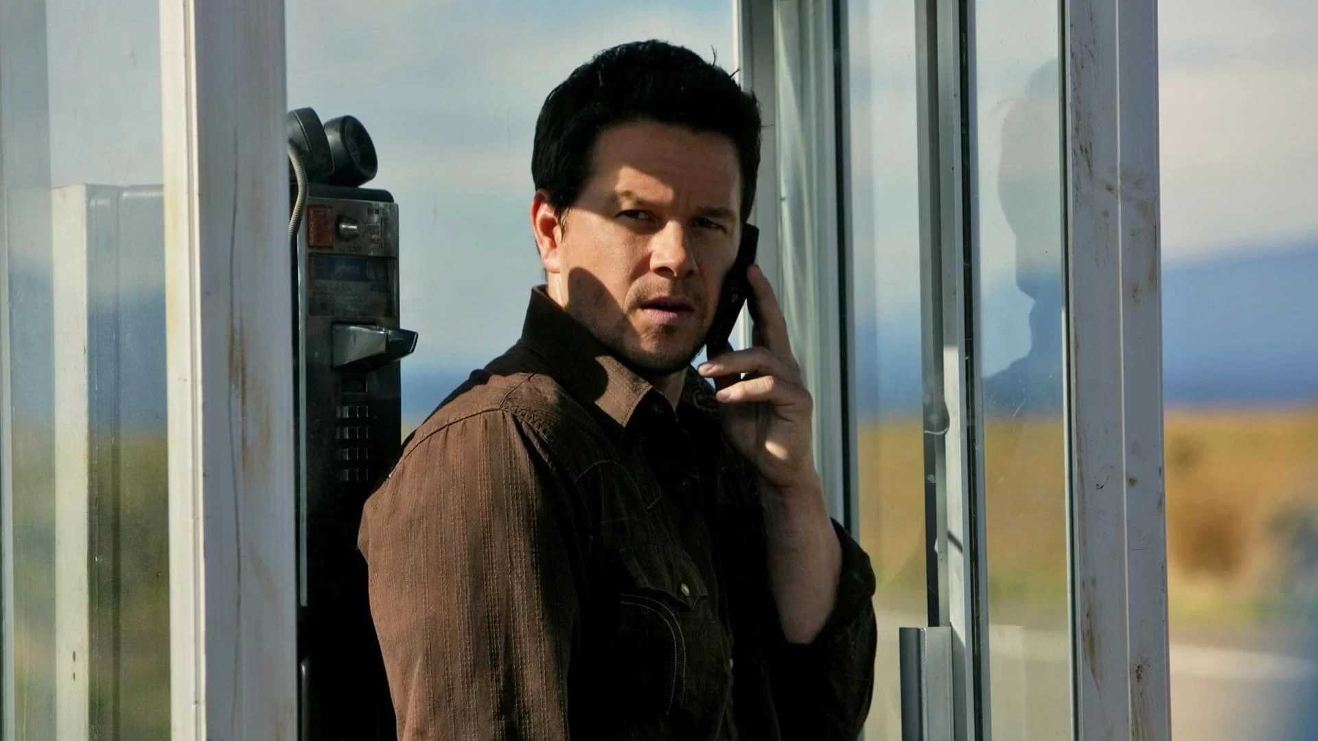 A man stands in a phone booth, speaking on a cellphone. He wears a brown jacket, and the booth is situated in a rural setting.