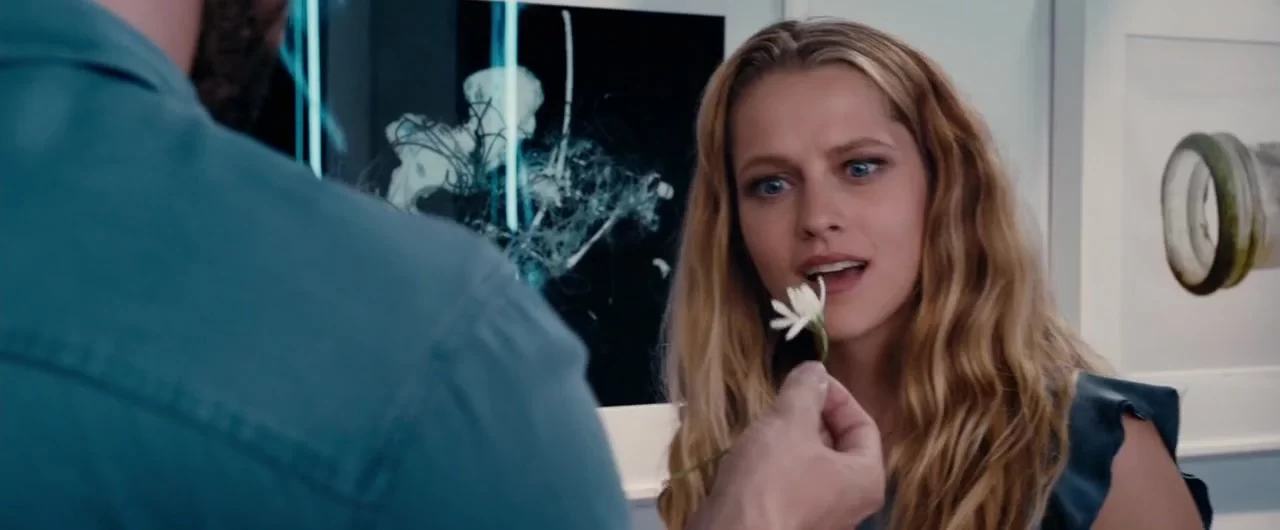 Person holding a white flower in front of a woman with long blonde hair, who is gazing at it intently. Abstract art and a large jar are in the background.