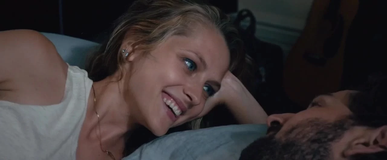 A woman lying on a bed, smiling while looking at a man beside her. Theres a guitar blurred in the background.