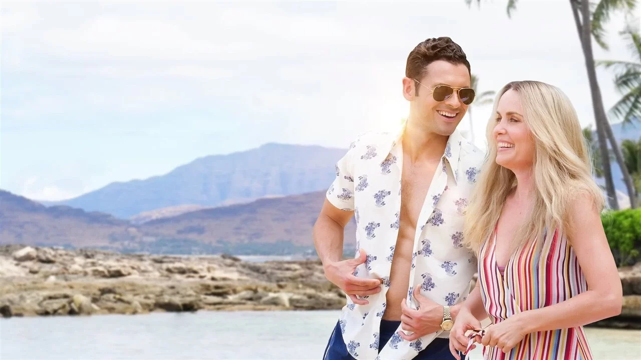 A man and woman in summer attire smile on a rocky beach with mountains in the background.