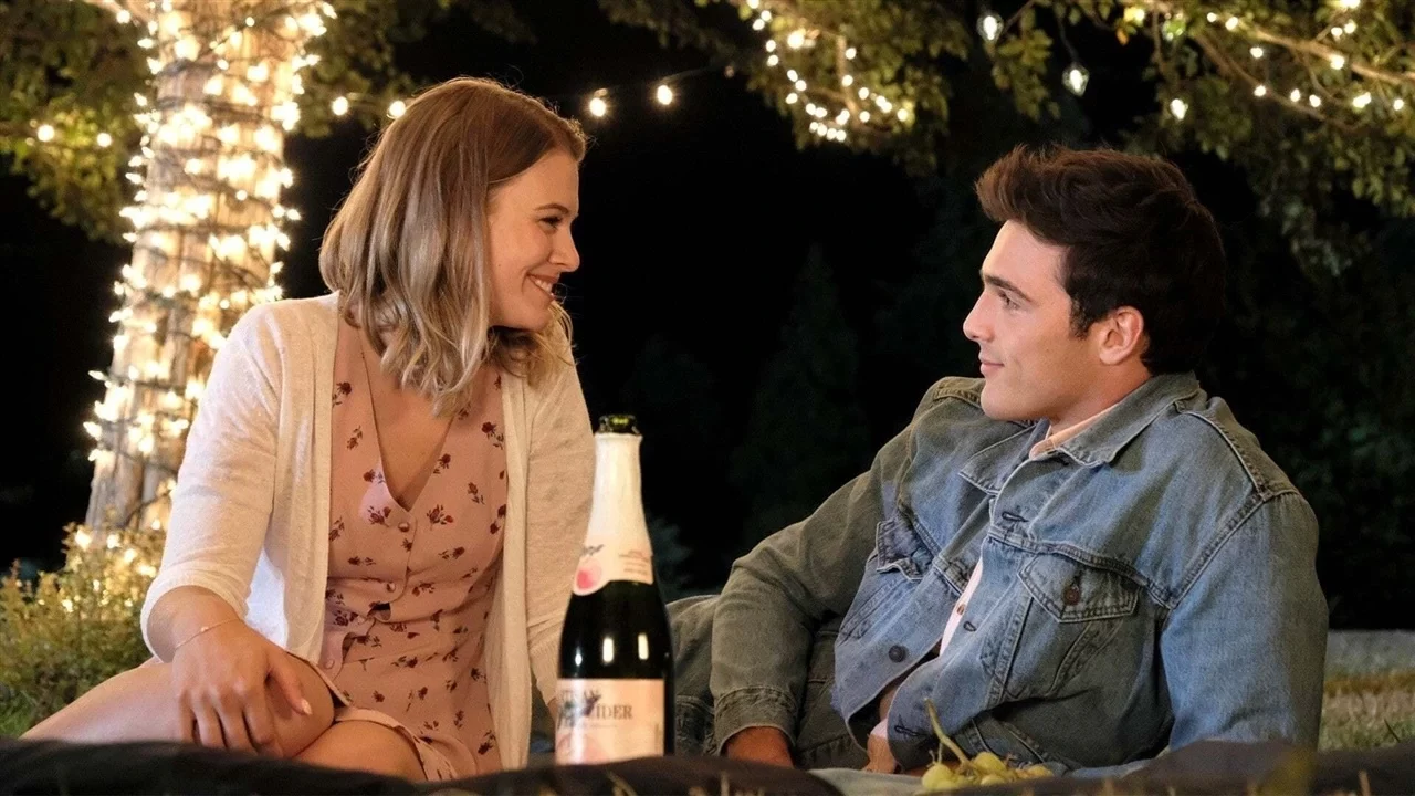 A woman and a man sit on a blanket under string lights at night, facing and smiling at each other. A bottle of sparkling wine is in front of them.