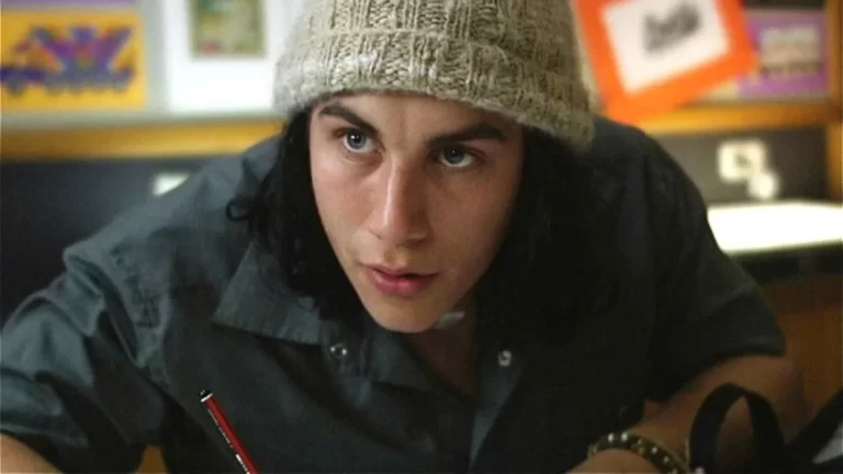Person wearing a knit hat and a gray shirt, sitting at a desk, writing with a pencil.
