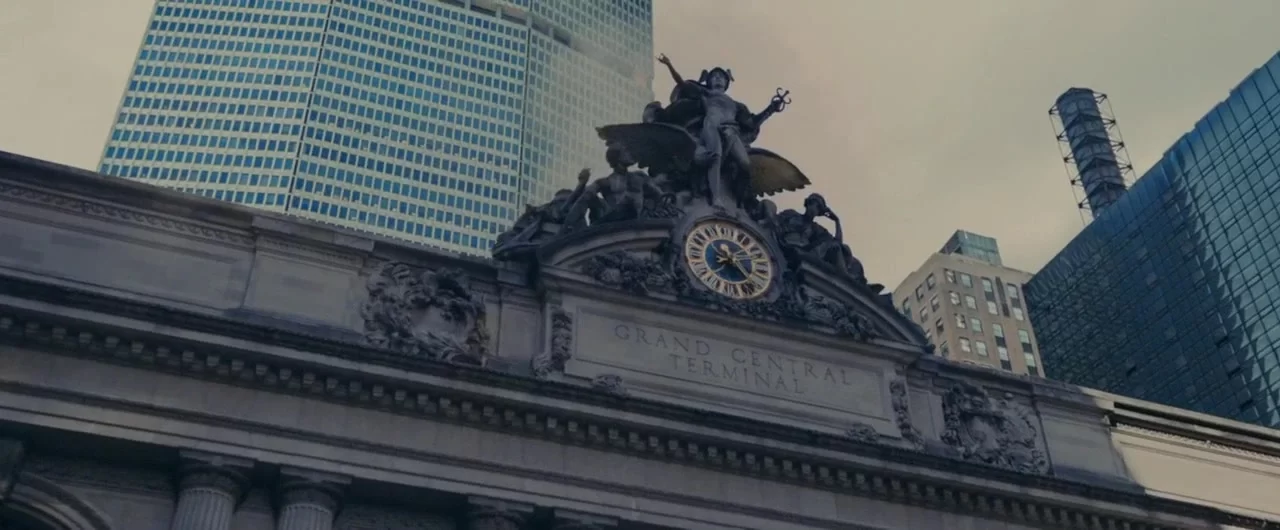 Facade of Grand Central Terminal showing its clock and sculptures with skyscrapers in the background under a cloudy sky.