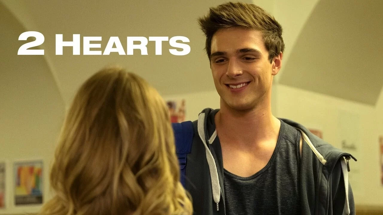 A young man with short brown hair smiles at a woman with long blonde hair. The text 2 Hearts is displayed in the upper left corner.