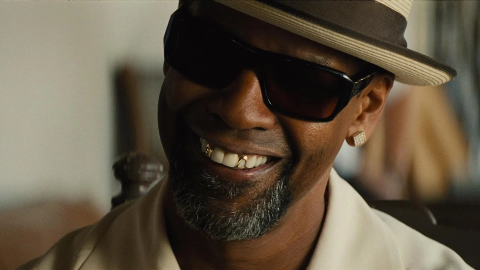 Denzel wearing sunglasses, a hat, and a beige shirt smiles, showing a gold tooth. They have a salt-and-pepper beard and an earring.