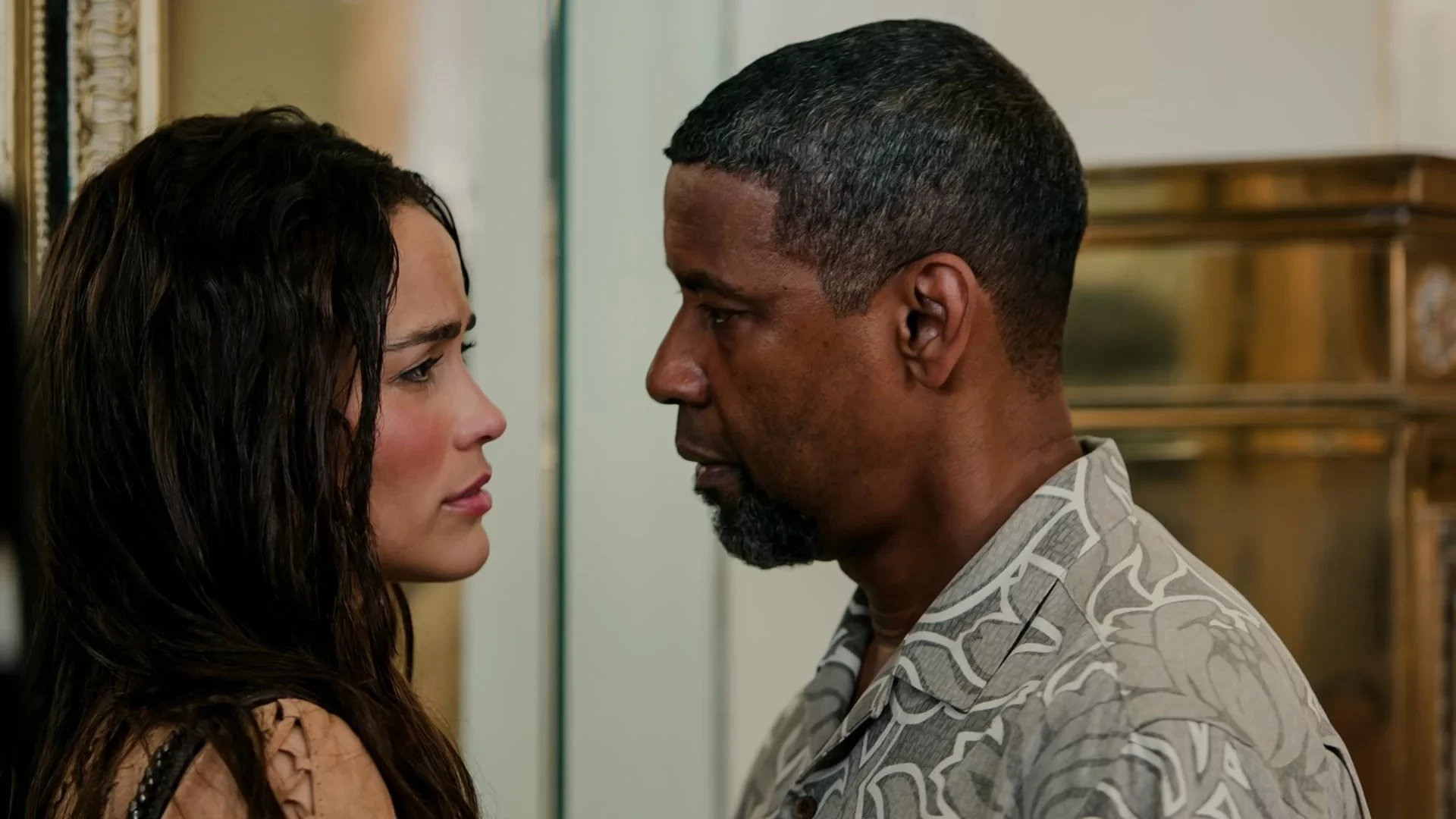 A man and woman stand close, facing each other in a tense moment. The woman has long, dark hair, and the man has short hair and a goatee. Both appear serious amidst a neutral indoor setting.