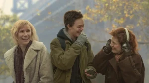 Three people in winter clothing stand outside. The person on the left is smiling, the person in the middle is laughing with their hand near their face, and the person on the right is adjusting or wearing large headphones. Trees are in the background.