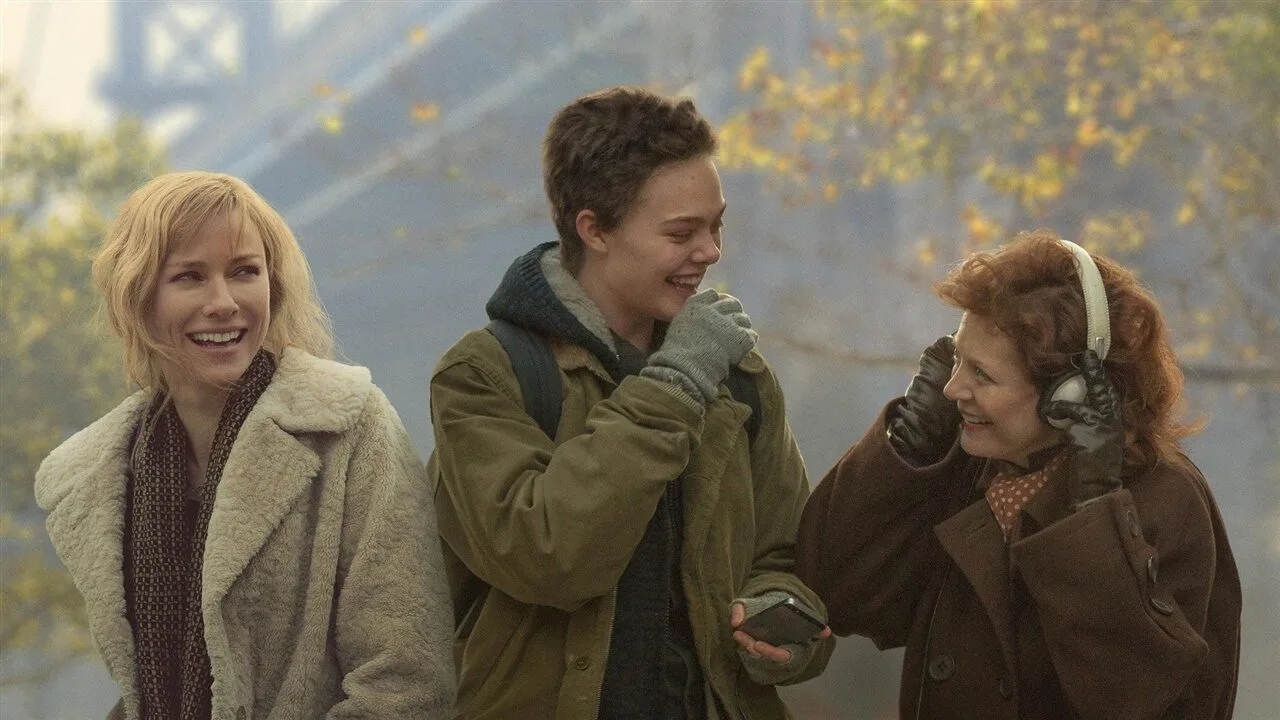 Three people in winter clothing stand outside. The person on the left is smiling, the person in the middle is laughing with their hand near their face, and the person on the right is adjusting or wearing large headphones. Trees are in the background.